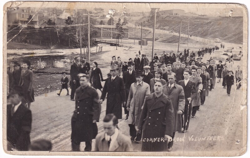 The image is a historical black-and-white photograph showing a long procession of people, primarily men, walking along a rural road. They are walking in an orderly line, and their expressions range from serious to neutral. In the background, there is a visible small river or creek with a few trees lining the banks. On the left side, there are residential buildings on a sloped landscape. The photograph shows signs of aging, with visible creases and worn edges.