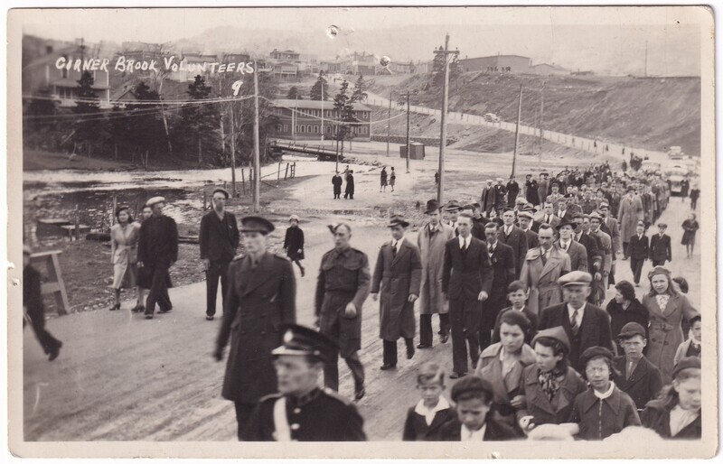 The black and white image shows a large group of people walking down a rural road. The road is muddy and flanked by a hilly landscape. In the background, there are several buildings, some partially obscured by trees. A bridge crosses a stream to the left. The people in the procession appear to be of different ages, from children to adults.