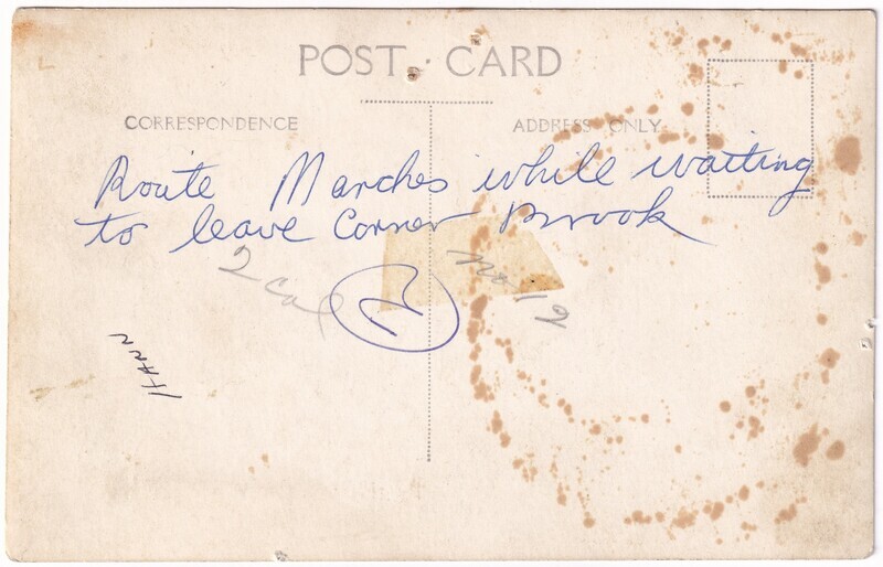 The image is of the worn and aged back of a photograph, featuring several brown stains and spots scattered across its surface. At the top, centered, is the printed text "POST CARD" in large capital letters. Below this, there are two sections labeled "CORRESPONDENCE" on the left and "ADDRESS ONLY" on the right, with a vertical dotted line running between them. Handwritten in blue ink is the text “Route Marches while waiting to leave Corner Brook.” Additional smaller and fainter handwriting is visible, including "Hann" written vertically in black ink, and an old piece of brown tape marks the center.