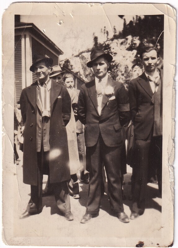 The black-and-white photograph depicts a group of men outdoors, dressed in formal attire. In the foreground, three men are prominently standing. The man on the left wears a long overcoat, a hat, and scarf draped around his neck. The middle man sports a suit jacket, tie, and a boutonnière on his lapel, with a hat tilted on his head. The man on the right also wears a suit and tie. Behind them, partially obscured, are other individuals similarly dressed. The backdrop consists of a building with vertical siding and a rocky landscape with trees. The photo's edges are worn and slightly damaged.