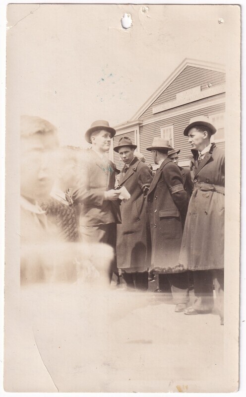 The black and white photograph features a group of men wearing long overcoats and hats standing outdoors in front of a wooden building. The building in the background has horizontal siding and a gable roof. The photo is slightly faded, with a hazy foreground where the figures are partially obscured.