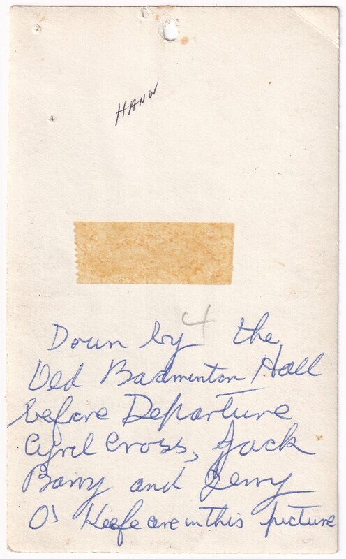 The image shows the back of a worn photograph featuring handwritten text in blue ink. There is a small hole near the top and some discoloration and wear around the edges. There is a piece of stained tape affixed near the center. The text "Down by the Old Badminton Hall before Departure Cyril Cross, Jack Barry and Jerry O'Keefe are in this picture" is handwritten in a cursive style in blue ink. The surface appears aged, with slight stains and marks.