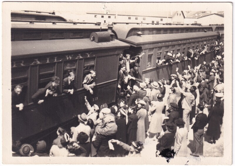 The black-and-white photograph shows a bustling scene at a railway station. Two adjacent train cars are filled with passengers leaning out of the windows to interact with a large, densely packed crowd of men, women, and children on the platform. Many in the crowd are reaching up towards the passengers, waving or holding out hands. The train cars are dark in color, with visible windows, and one of them displays partially visible text: "RAILWAY" and the number "18."