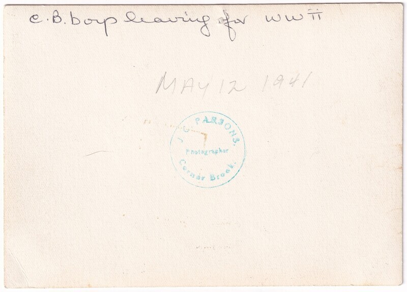 The image shows the back of an aged photograph featuring handwritten text and a blue stamp. The handwritten inscription along the top left reads, "C.B. boys leaving for WWII." Below, it reads, "MAY 12 1941" in capital letters. In the lower central part of the image, there is a round blue stamp with the inscription "J.C. Parsons, Photographer, Corner Brook."