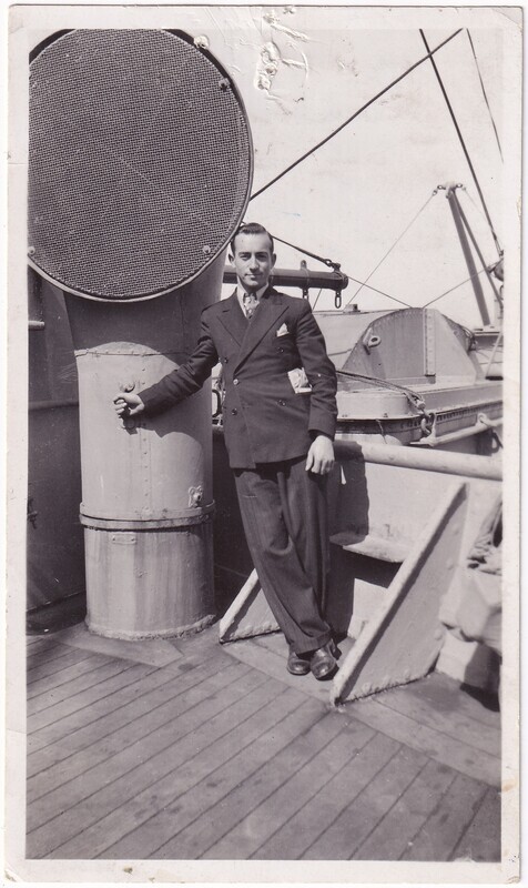 The image is a black and white photograph of a man in a suit leaning against the a large cylindrical ventilation duct on a ship's deck. The man is dressed in a double-breasted suit with a tie and pocket square. The deck is wooden, and the background shows metal structures and cables.