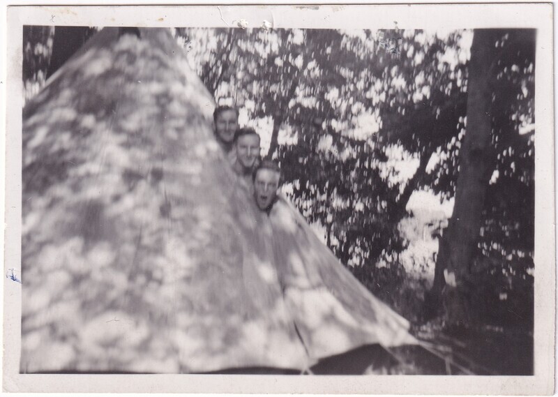 The black and white photograph depicts three people partially emerging from inside a conical tent. The tent is on the left side of the image and reflects dappled sunlight filtering through leaves. The three individuals are lined vertically. They are surrounded by a densely wooded area. The photograph has a slight blurriness.