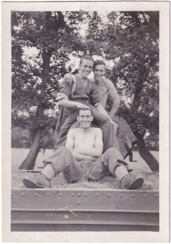 The image is a black and white photograph depicting three young men outdoors. The man in the foreground is seated cross-legged on the ground and shirtless. Behind him, two other men stand, with the one on the left slightly leaning forward with his right hand rested on the seated man's head. Both of the standing men are partially clothed in loose-fitting, casual attire. The background shows large trees with abundant foliage.