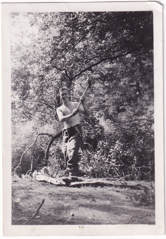 The black and white image depicts a man standing outdoors in a wooded area. He is shirtless and appears to be smiling while holding an axe with both hands. The man is wearing pants and his posture suggests he might be chopping wood. The background consists of dense foliage, with tree branches and leaves creating a canopy above him.