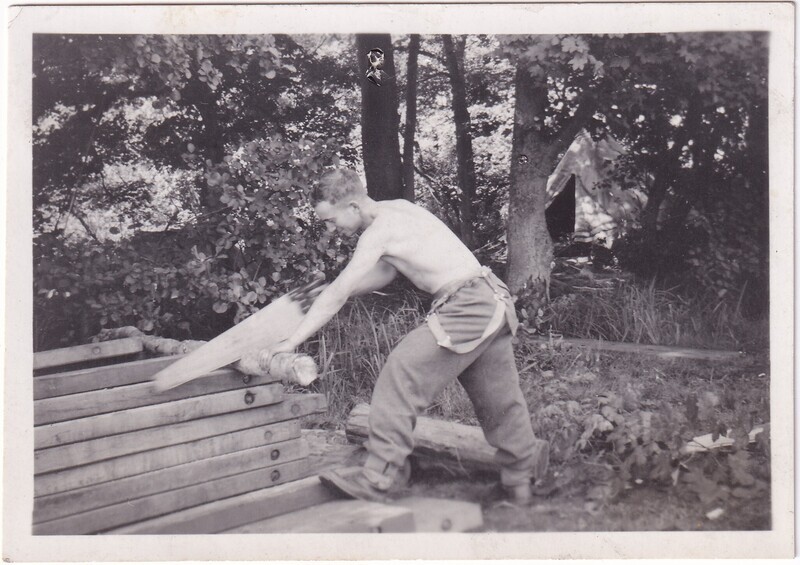 The black-and-white image shows a man engaged in manual work outdoors. He appears to be shirtless and is wearing loose pants. The man is using a saw to cut through a piece of wood, which is positioned on a stack of similar beams. The scene is set in a wooded area with trees and dense foliage in the background. There is a tent partially visible among the trees