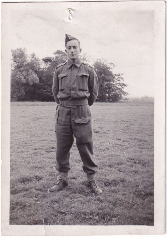 The image is a black-and-white photograph of a person in military uniform standing on a grassy field. The individual is facing the camera with hands clasped behind their back, wearing a buttoned-up jacket with epaulets and two breast pockets. The trousers are tucked into sturdy boots. A side cap is worn at an angle on the head. In the background, there are trees lining the horizon, and the sky is overcast.