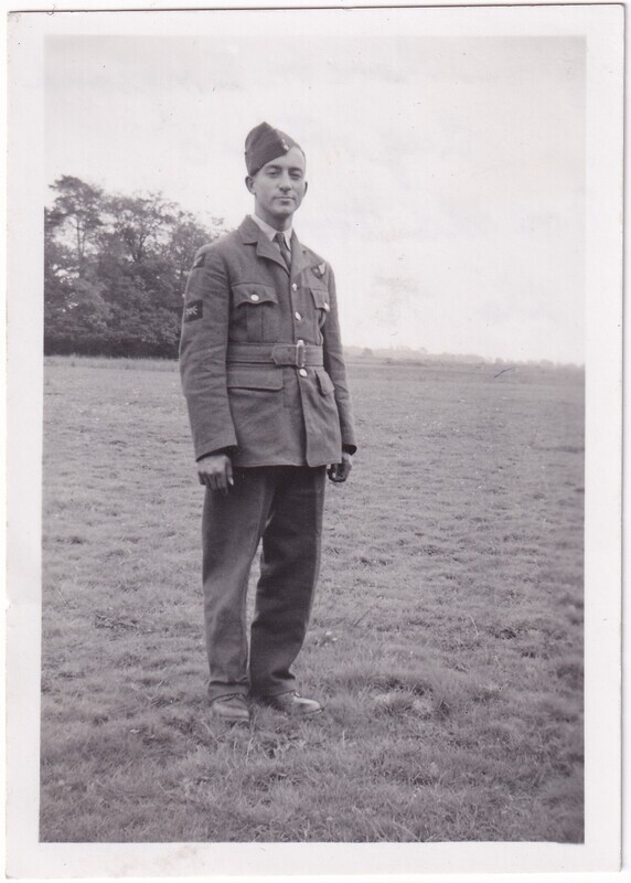 The black and white photograph depicts a man standing outdoors on a grassy field. He is wearing a military uniform, consisting of a buttoned jacket, belted at the waist, and matching trousers. A cap is tilted on his head. The background shows a line of trees on the left, with an open field stretching to the right under a cloudy sky. The man is standing with his hands by his sides, looking directly at the camera