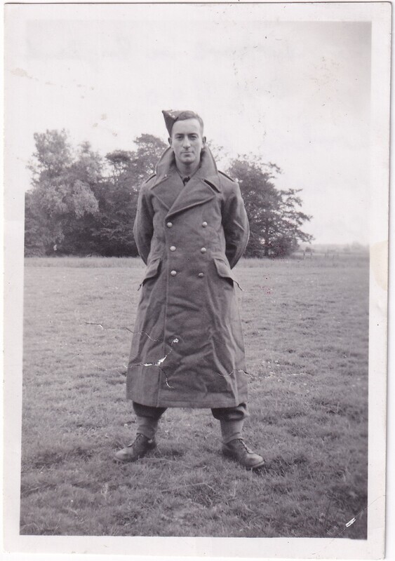 The black-and-white photograph features a person standing outdoors in an open field with a backdrop of trees. The individual is wearing a long, double-breasted, military-style overcoat with prominent lapels and visible buttons. The coat reaches down to their shins. They have their hands tucked behind their back and are wearing a side cap tilted to the side of their head. The person is looking directly at the camera, standing on grass, with trees visible in the distance under a cloudy sky.