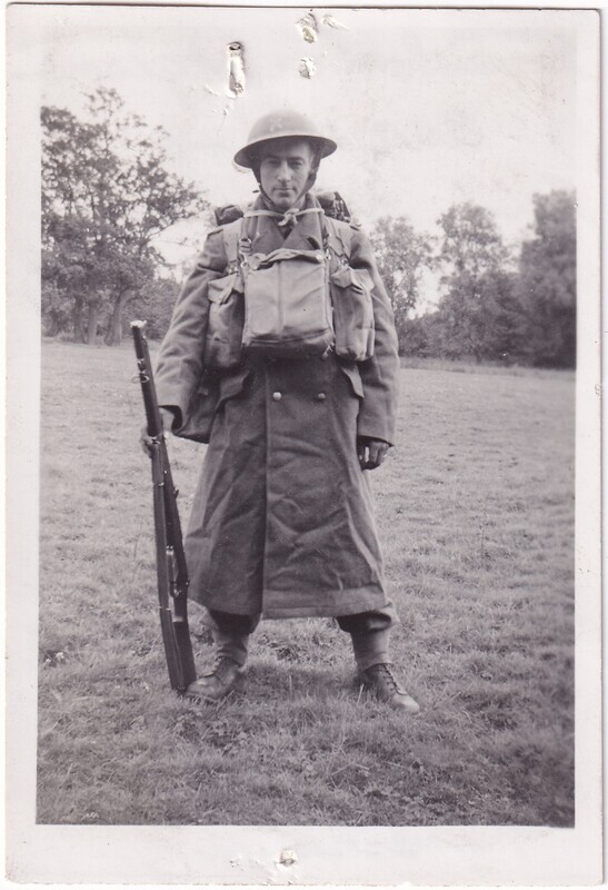 The black-and-white photograph features a soldier standing outdoors on a grassy field. He is dressed in a heavy, long military coat with large buttons and a metal helmet. A large, square backpack is secured to his front with straps over his shoulders. He holds a rifle vertically in his right hand, resting on the ground. Behind him, there are trees with dense foliage