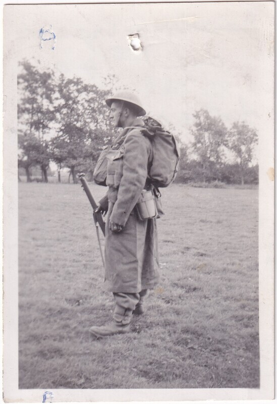 The image is a black and white photograph of a soldier standing in profile on an open field. The soldier is dressed in a heavy coat and wearing a helmet. He carries a large backpack and several pouches attached to his belt. In his right hand, he holds a rifle with the butt of the gun resting on the ground. The field is grassy, extending to a line of trees in the background