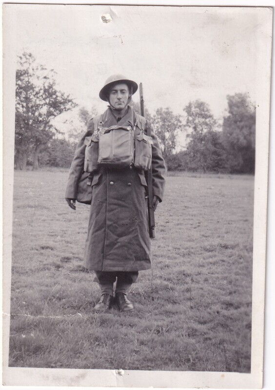 The image is a black and white photograph depicting a soldier standing in a grassy field. The soldier is wearing a long coat and a helmet, with a large backpack strapped to his front and additional gear on his back. He holds a rifle upright by his side. The background features a line of trees