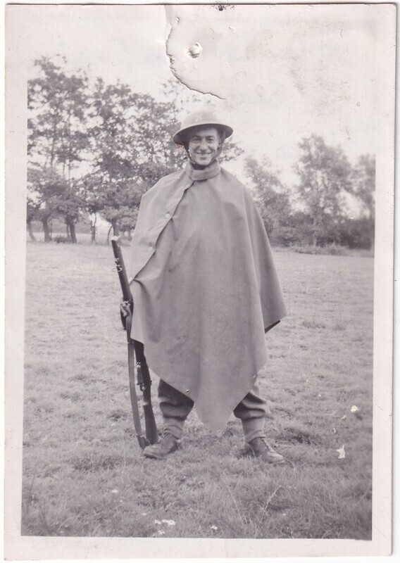 The black and white photograph features a person standing in an open grassy field. The individual is wearing a military-style helmet and a large cape that covers most of their body. The person is smiling and holding a rifle vertically in one hand. In the background, there are several trees with dense foliage