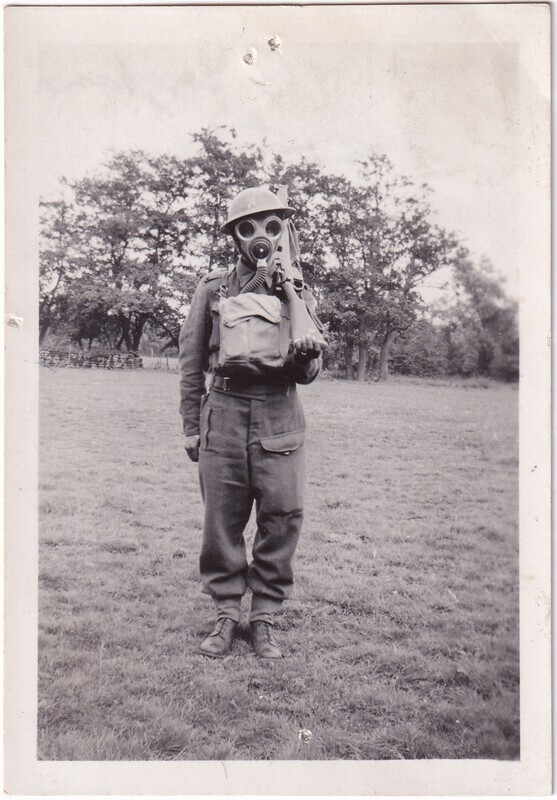 The black and white photograph depicts a person dressed in military attire, standing on a grassy field. The individual is wearing a gas mask with large circular eye lenses and a protruding filter, alongside a helmet. A uniform, consisting of a buttoned jacket and trousers, is worn along with sturdy boots. The background shows a line of trees