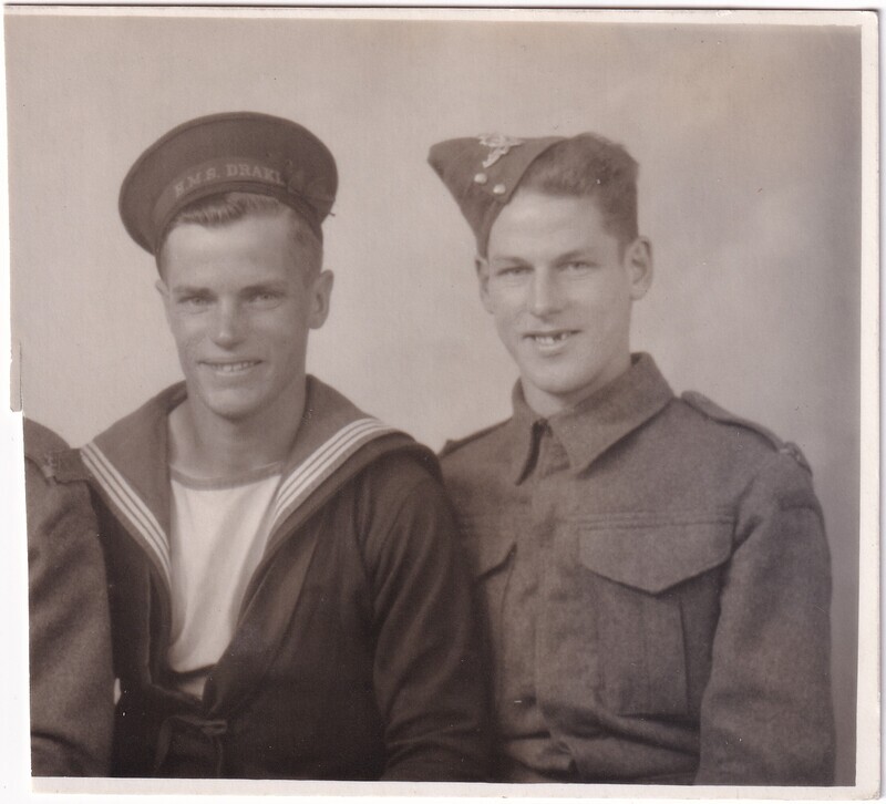 The image is a black and white photograph of two men in military uniforms, seated side by side. On the left, a man is dressed in a naval uniform, identifiable by the sailor's hat marked "H.M.S. DRAKE" and a traditional striped collar with a tied front. His expression is neutral, with a slight smile. On the right, the second man is wearing a military uniform with a high-collared jacket featuring two pockets on the chest. He is also wearing a cap with a badge. The background is plain