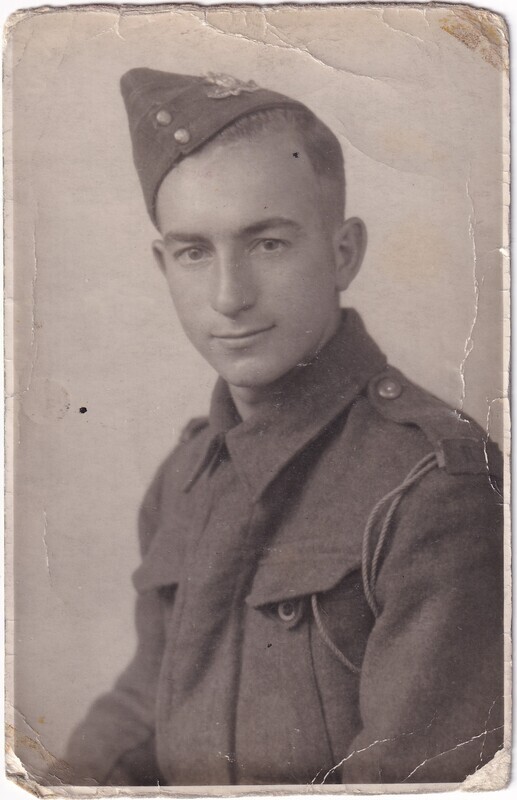 The image is a sepia-toned vintage portrait photograph of a man dressed in military uniform. He is looking directly at the camera with a slight smile. The man is wearing a peaked cap with insignia, which includes a badge on the front. His uniform includes a high-collared jacket with prominent buttons on each shoulder. The image appears old, with visible creases and worn edges, typical of aged photographs. The background is plain, highlighting the subject.