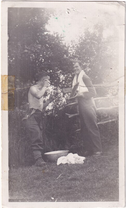 The black and white photograph depicts two men outdoors, set against a background of dense foliage. The man on the left is shirtless, wearing trousers and boots, and appears to be shaving with a straight razor, holding a mirror near his face. Beside him on the grass is a basin and a pile of cloth or towels. The man on the right is standing, dressed in dark trousers and a sweater, with a towel draped over his shoulder, smiling and holding what appears to be a camera or another object. Behind them is a rustic wooden fence, and the ground is covered with grass.