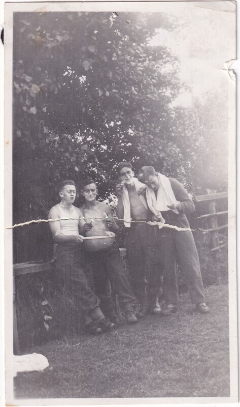 The black and white photograph depicts four men outdoors, possibly during a playful moment of shaving or washing up. They are standing close together on a grassy area bordered by dense foliage and wooden fencing. Each man is partially dressed, wearing trousers and either undershirts or no shirt at all. The first man on the left holds a metal bowl, while the second and third men have shaving cream on their faces, one holding a straight razor. The fourth man laughs with a towel draped around his neck. The image shows signs of wear, with creases and scratches visible.