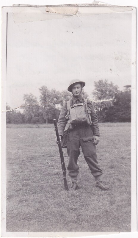 The image is a black and white photograph depicting a soldier standing in a grassy field. He is wearing a military uniform with a helmet and is carrying a rifle in his right hand, which is pointed downwards. His uniform includes a woolen overcoat, pants with large pockets, and boots. He also has a large backpack strapped to his chest. In the background, there are trees at a distance, and the sky appears overcast, suggesting the photograph was taken during the day. The photograph shows some signs of aging, such as creases and a slight tear at the top edge.