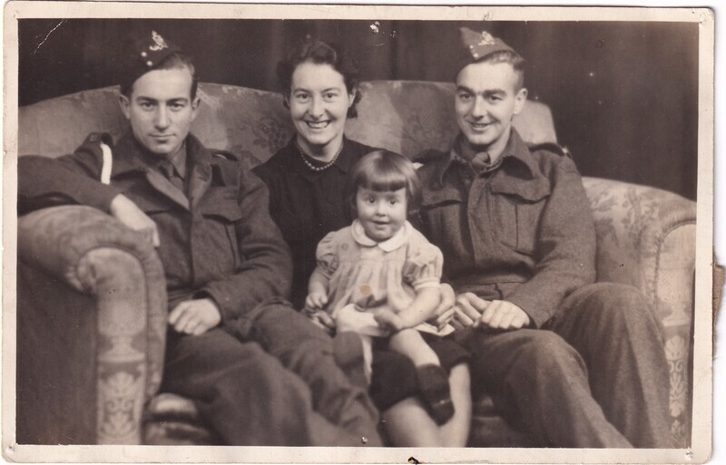 This is a black and white photograph showing a group of four individuals sitting together on a patterned couch. On the left, a man in a military uniform with a hat is seated. Next to him, a woman in a dark dress with a pearl necklace sits, smiling. In the center front, a young child in a light-colored dress with short hair is perched, looking at the camera. To the right, another man in a similar military uniform and hat is seated, also smiling. The backdrop is plain, possibly a curtain, creating a focused composition around the individuals.